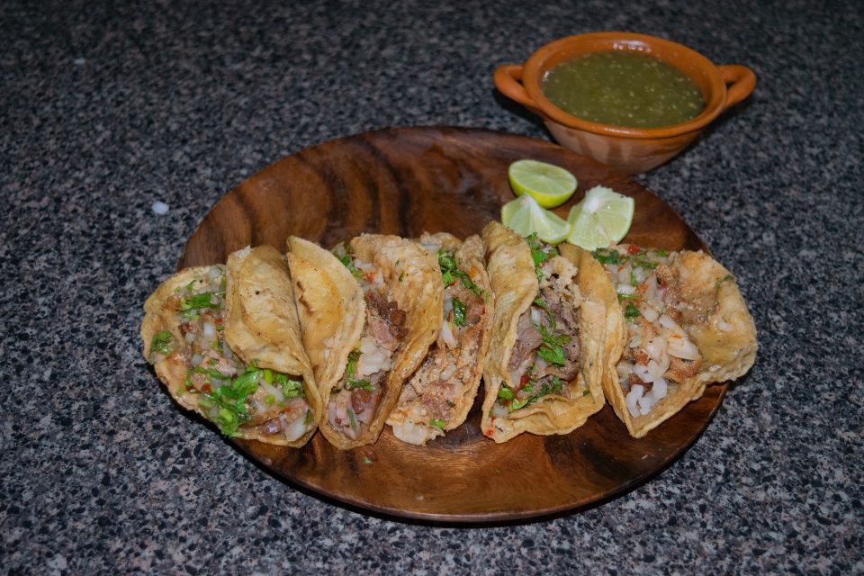 A wooden plate with street tacos with a side of salsa verde.